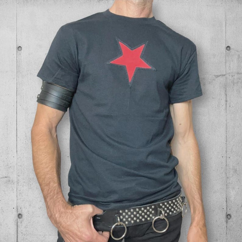A black t-shirt with a red star on the front. The t-shirt is worn with a black leather bracelet on the left wrist and a black studded belt with silver rings.