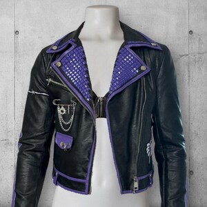 May include: Black leather jacket with purple accents, including studded lapels and trim. Features a zipper closure, pockets, and decorative chain and safety pin details. The jacket is displayed on a mannequin.