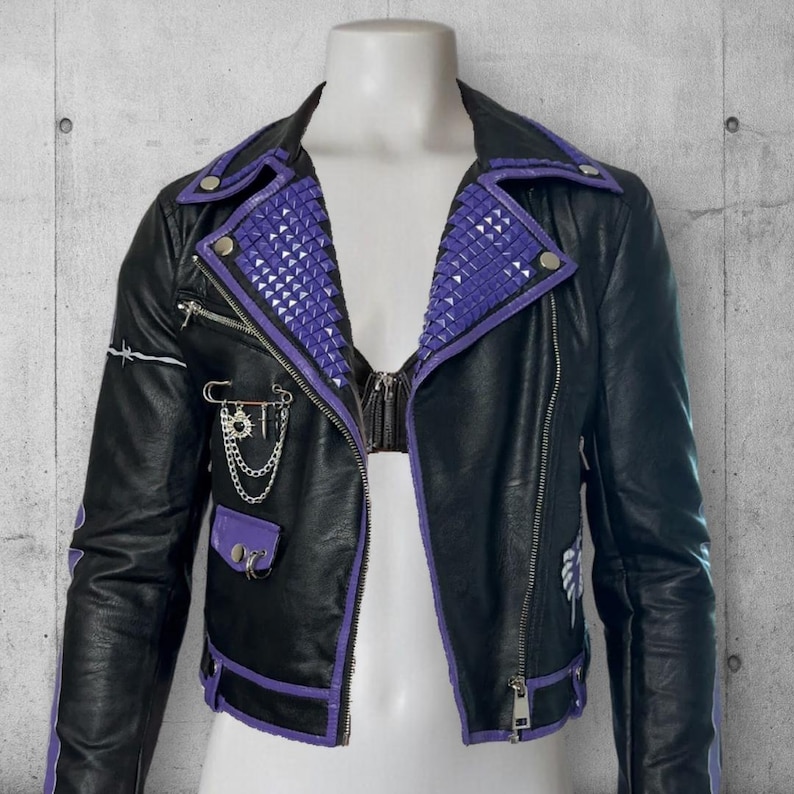 May include: Black leather jacket with purple accents, including studded lapels and trim. Features a zipper closure, pockets, and decorative chain and safety pin details. The jacket is displayed on a mannequin.