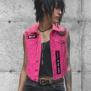May include: A bright pink denim vest with silver studs and a faux fur collar. The vest features patches that read "RIOT" and "enemy of the state". The vest has a classic button-up design and two front pockets. A punk rock fashion statement.