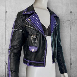 May include: Black faux leather jacket with purple accents, including studded lapels and sleeve stripes. Features a zippered front, multiple pockets, and decorative chain and safety pin details. A bold, edgy fashion statement.