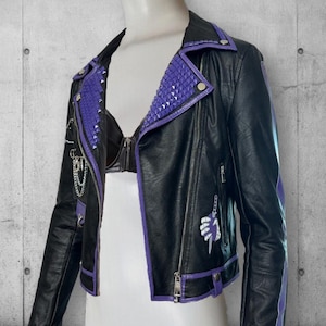 May include: Black faux leather jacket with purple trim and studded accents. The jacket features a zipper closure, a small pocket, and a decorative chain. A stylized hand graphic is on the front. The jacket is displayed on a mannequin.
