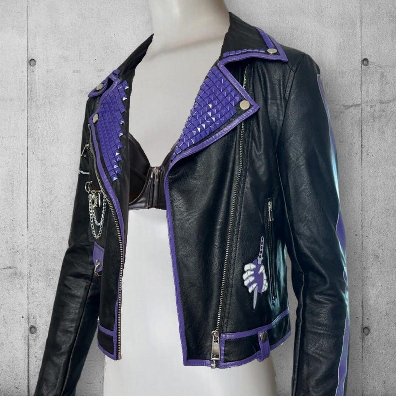 May include: Black faux leather jacket with purple trim and studded accents. The jacket features a zipper closure, a small pocket, and a decorative chain. A stylized hand graphic is on the front. The jacket is displayed on a mannequin.