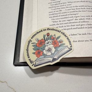 May include: A sticker with the text "I get emotionally attached to fictional characters" in a semi-circle around a floral design. The design features an open book with flowers in shades of orange, white, and teal.