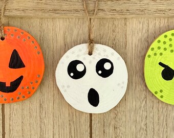 May include: Three round wooden ornaments painted with Halloween faces. The first ornament is orange with a jack-o'-lantern face. The second ornament is white with a ghost face. The third ornament is green with a zombie face.
