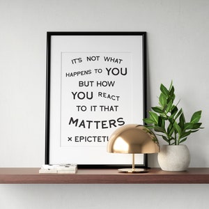 May include: A black framed print with a white background and black text that reads "IT'S NOT WHAT HAPPENS TO YOU BUT HOW YOU REACT TO IT THAT MATTERS × EPICTETI". A gold dome lamp sits on a wooden shelf with a potted green plant.