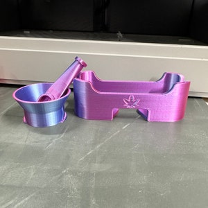 Puede incluir: Un conjunto de tres herramientas impresas en 3D de color morado y azul para moler hierbas. Las herramientas incluyen un mortero y un pilón, y una bandeja pequeña con un diseño de hoja.