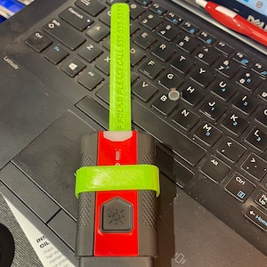 Könnte beinhalten: Ein grün-rotes Gerät mit einem leuchtend gelben Band. Das Gerät hat einen Knopf mit einem Sonnensymbol und der Text "IF FOUND PLEASE CALL 555 555 5555" ist auf dem Band aufgedruckt. Das Gerät liegt auf einer schwarzen Laptop-Tastatur.