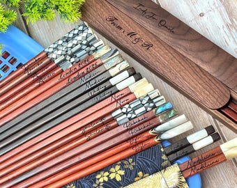 Könnte beinhalten: Eine Sammlung von Essstäbchen aus Holz mit eingravierten Namen auf den Griffen, verziert mit Einlagen aus Perlmutt, Abalone und metallischen Mustern. Das Set enthält ein Aufbewahrungsetui aus dunklem Walnussholz mit der Gravur From M & R.
