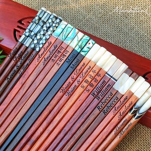 May include: A collection of wooden chopsticks featuring various inlaid decorative tops, including mother-of-pearl and abalone shell patterns. Each pair is engraved with names such as Peter, Amelinda, Mosi, Brian & Laura, William, Rebecca, Amilone, and Akirami.