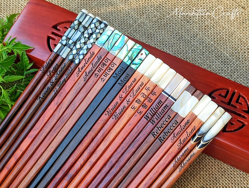 May include: A collection of wooden chopsticks featuring various inlaid decorative tops, including mother-of-pearl and abalone shell patterns. Each pair is engraved with names such as Peter, Amelinda, Mosi, Brian & Laura, William, Rebecca, Amilone, and Akirami.