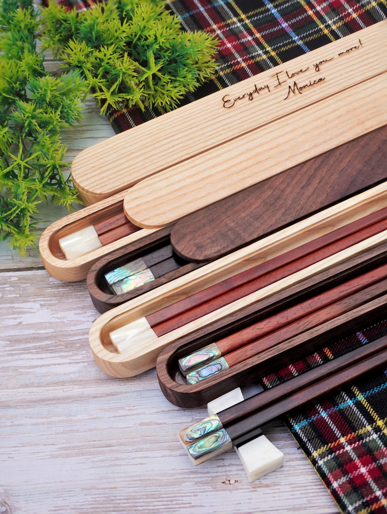 May include: A collection of wooden chopstick sets in sliding cases. The top case is light wood with the engraved text "Everyday I love you more! Monica". Other cases are dark wood, containing chopsticks with iridescent shell inlays.