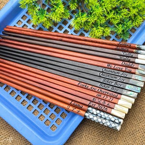 May include: A collection of wooden chopsticks in varying shades of brown and black, each featuring unique laser-engraved text including names like James, Liana, and phrases such as We Love You and Forks Are Overrated, with decorative inlaid ends.
