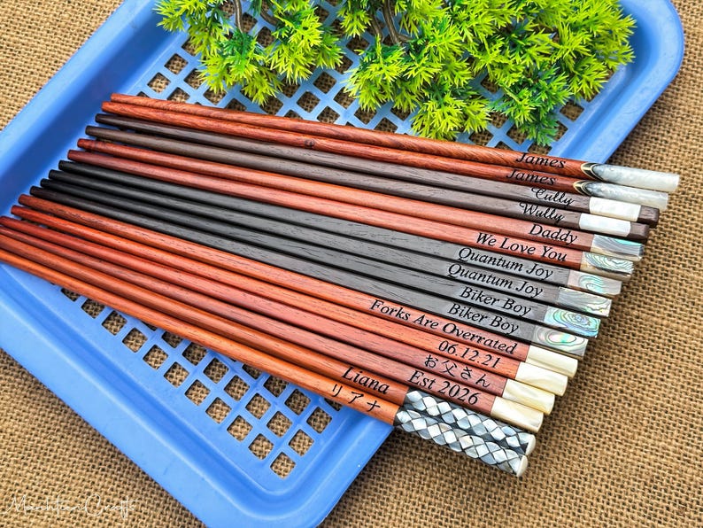 May include: A collection of wooden chopsticks in varying shades of brown and black, each featuring unique laser-engraved text including names like James, Liana, and phrases such as We Love You and Forks Are Overrated, with decorative inlaid ends.