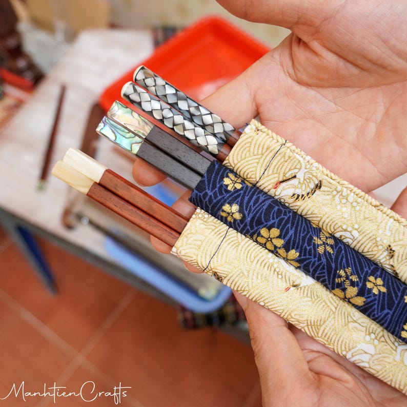 May include: Three pairs of chopsticks with distinct handles: one pair with light wood, one with dark wood and iridescent abalone inlay, and one with dark wood and a woven silver-toned pattern. Each pair is housed in a fabric sleeve with gold and blue patterns.