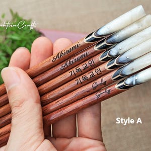 May include: A set of six wooden chopsticks with smooth brown grain, featuring personalized names and Korean text engraved on the shafts. The handles are capped with a polished, marbled white and black horn-like material, accented with a small silver stud.