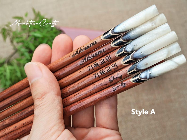 May include: A set of six wooden chopsticks with smooth brown grain, featuring personalized names and Korean text engraved on the shafts. The handles are capped with a polished, marbled white and black horn-like material, accented with a small silver stud.