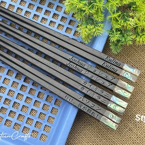 May include: A set of five dark wood chopsticks featuring iridescent abalone shell inlays at the ends. Each pair is engraved with names and phrases including Joshua, Eulina, and Japanese text. The chopsticks are arranged diagonally on a blue plastic tray.