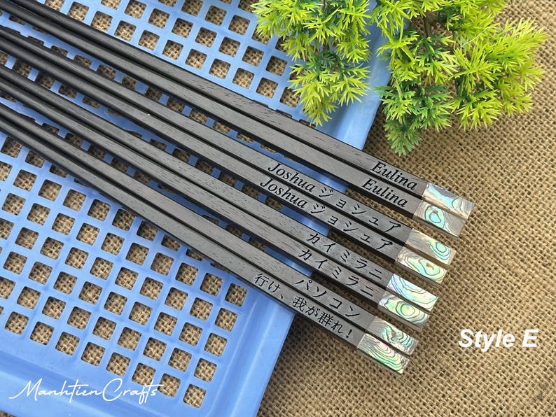 May include: A set of five dark wood chopsticks featuring iridescent abalone shell inlays at the ends. Each pair is engraved with names and phrases including Joshua, Eulina, and Japanese text. The chopsticks are arranged diagonally on a blue plastic tray.