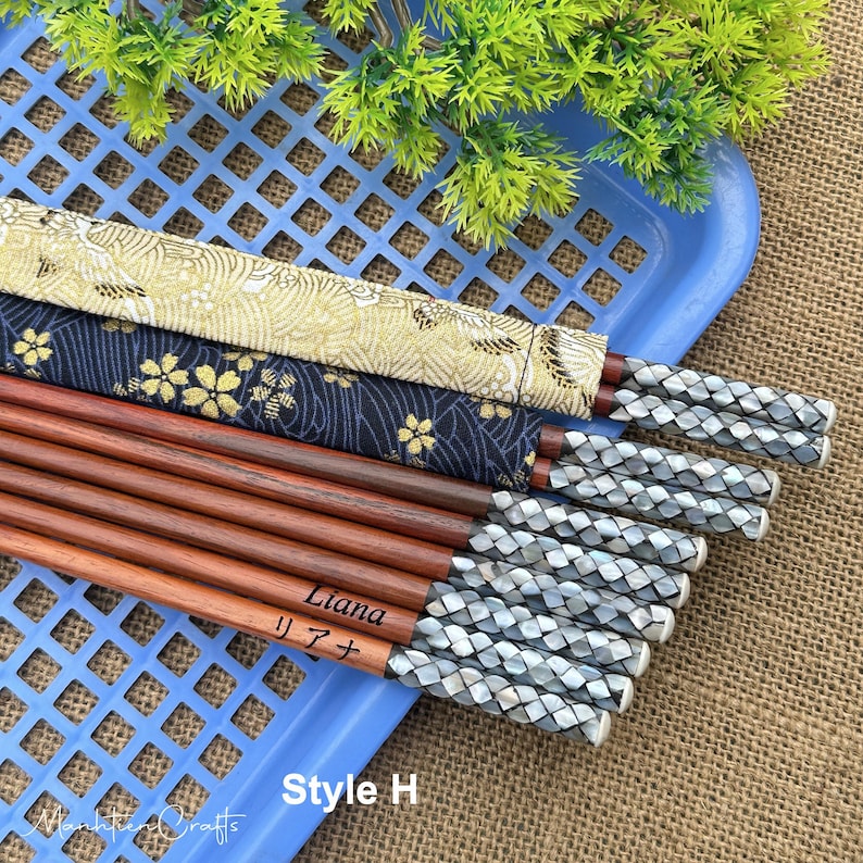 May include: A set of wooden chopsticks with braided silver-toned ends. Some chopsticks feature fabric sleeves with gold and navy floral patterns. One chopstick is engraved with the name Liana in English and Japanese characters. The text Style H is visible.