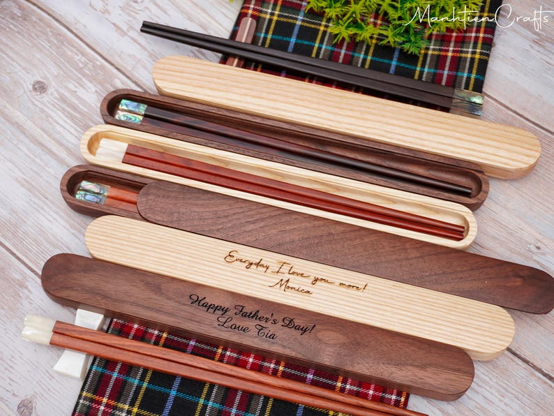 May include: A collection of wooden chopstick sets with matching cases. Some cases are light wood, others are dark wood. Two cases feature engraved text: "Everyday I love you more! Monica" and "Happy Father's Day! Love Tia." The chopsticks have inlaid details.