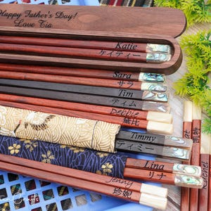 May include: A collection of wooden chopsticks with personalized names in English and Japanese, featuring iridescent shell inlays at the ends. Some are stored in a dark wood box engraved with "Happy Father's Day! Love Tia" and others in fabric sleeves.