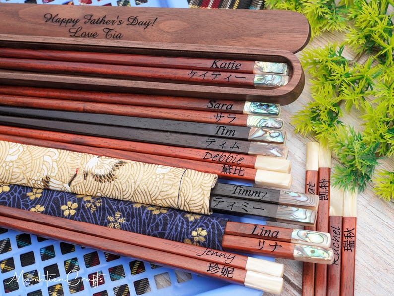 May include: A collection of wooden chopsticks with personalized names in English and Japanese, featuring iridescent shell inlays at the ends. Some are stored in a dark wood box engraved with "Happy Father's Day! Love Tia" and others in fabric sleeves.