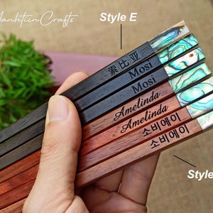 May include: A set of six wooden chopsticks featuring iridescent abalone shell inlays at the ends. The chopsticks are engraved with various names and text including Mosi, Amelinda, and characters. The set is labeled with Style E and Style F.