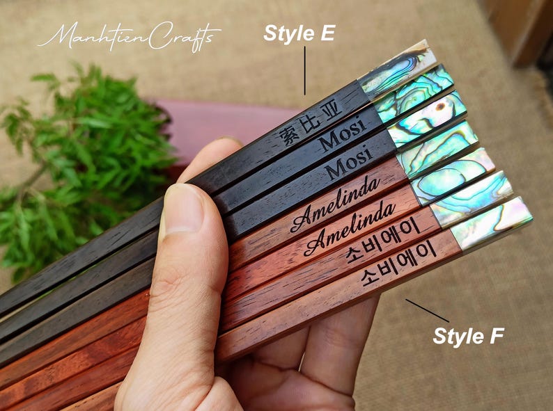 May include: A set of six wooden chopsticks featuring iridescent abalone shell inlays at the ends. The chopsticks are engraved with various names and text including Mosi, Amelinda, and characters. The set is labeled with Style E and Style F.