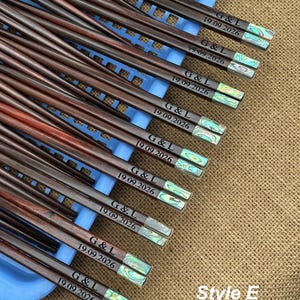 May include: A collection of dark brown wooden chopsticks featuring iridescent abalone shell inlays at the ends. Each pair is engraved with the text G & L and the date 19.09.2026. The chopsticks are arranged on a light blue plastic tray.