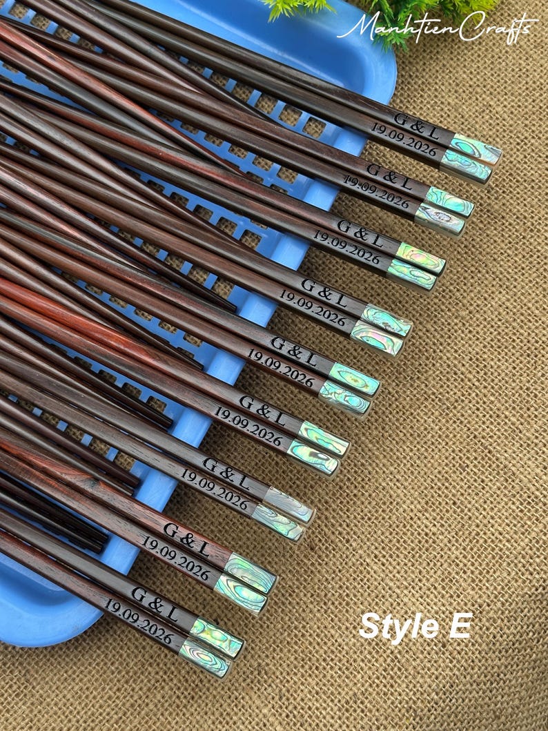 May include: A collection of dark brown wooden chopsticks featuring iridescent abalone shell inlays at the ends. Each pair is engraved with the text G & L and the date 19.09.2026. The chopsticks are arranged on a light blue plastic tray.