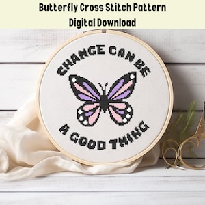 May include: A cross stitch pattern featuring a purple and pink butterfly with black outlines and the text "Change can be a good thing". The pattern is displayed in a wooden embroidery hoop on a white wooden surface.