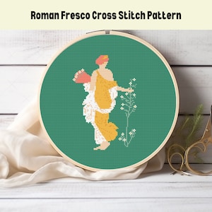 May include: A cross stitch pattern of a woman in a yellow dress holding a basket of flowers and standing next to a plant with white flowers. The pattern is on a green background and is framed in a wooden hoop.