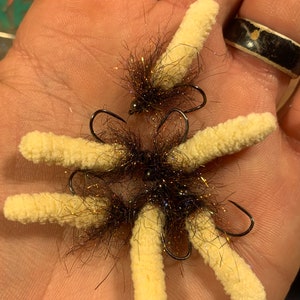 May include: A close-up of six brown and black fly fishing lures with white fuzzy bodies. The lures are tied to hooks and are arranged in a circle on a hand.