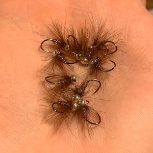 May include: A group of six brown and purple fly fishing lures with silver beads. The lures are tied on hooks and have feathers and fur.
