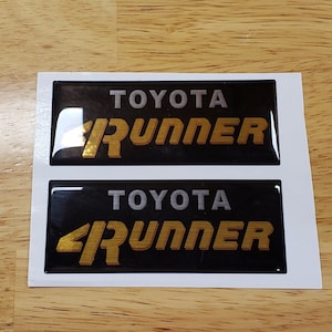 May include: Two black and silver "TOYOTA 4RUNNER" decals with a gold 3D effect.