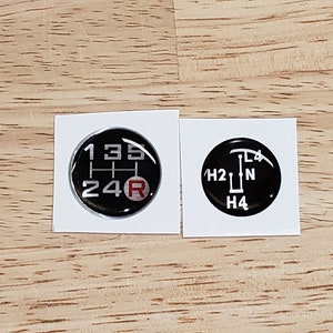 May include: Two black and white round stickers with a gear shift pattern. The first sticker shows a 5-speed manual transmission pattern with the numbers 1, 2, 3, 4, 5, and R. The second sticker shows a 4-wheel drive pattern with the letters H2, L4, N, and H4.