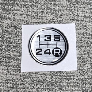 May include: A circular silver-toned gear shift pattern emblem featuring black engraved numbers 1, 3, 5 on the top row and 2, 4, R on the bottom row. The emblem has a brushed metallic finish with a polished silver border, presented on a square white backing.