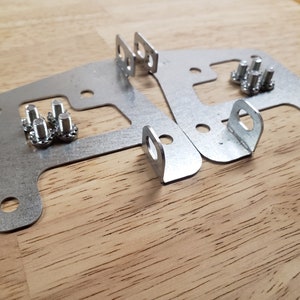 May include: Two silver metal brackets with multiple holes and a curved design, accompanied by several small silver screws. The brackets have a loop on one end. Hardware for construction or repair.