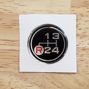 May include: A silver and black round sticker with a gear shift pattern. The numbers 1, 2, 3, and 4 are in silver, and the letter R is in red.