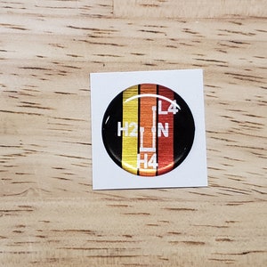May include: A round, black and gold sticker with a white gear shift pattern. The sticker has the letters H2, N, L4, and H4 printed on it.