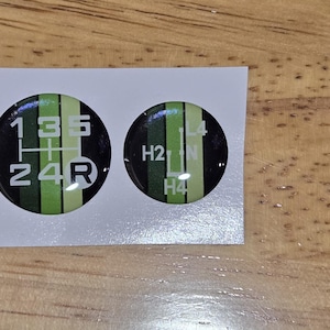 May include: Two circular stickers with a black background and green and white stripes. The larger sticker shows a gear shift pattern with numbers and letters. The smaller sticker has numbers and letters. Both stickers are on a white background.