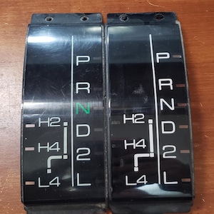 May include: Two black plastic automotive transmission shift indicator panels with white lettering. The left panel shows the letters "P", "R", "N", "D", "2", and "L" with a green "N". The right panel shows the letters "P", "R", "N", "D", "2", and "L". Both panels have the letters "H2", "H4", and "L4" on them.