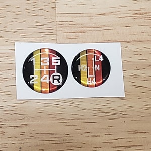 May include: Two round, black, and gold car gear shift knob stickers. The left sticker has the numbers 1, 2, 3, 4, 5, and R printed in white. The right sticker has the letters H2, N, L4, and H4 printed in white.