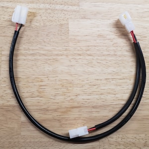 May include: A pair of black electrical wires with white connectors on each end. The wires are connected to each other and are lying on a wooden surface.