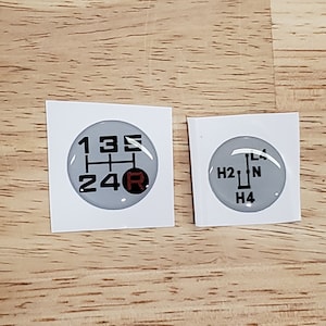 May include: Two white stickers with black and red gear shift patterns. The left sticker shows a 5-speed manual transmission pattern with the numbers 1, 2, 3, 4, 5, and R. The right sticker shows a 4-speed automatic transmission pattern with the letters H2, N, and H4.