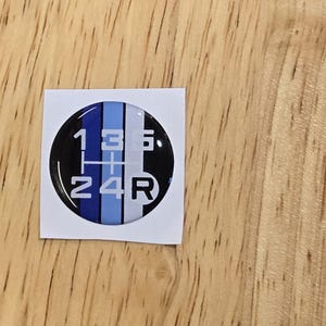 May include: A round, black and white sticker with blue stripes. The sticker has the numbers 1, 2, 3, 4, 5, and R printed on it.