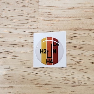 May include: A round, white, 3D sticker with a black gear shift pattern. The sticker has a red, orange, and yellow striped background. The gear shift pattern shows the letters 'H2', 'N', 'L4', and 'H4'.