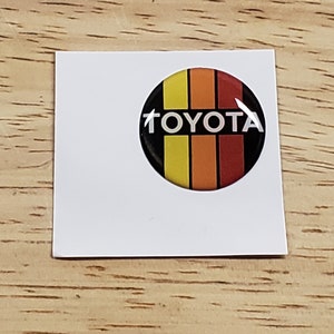 May include: A round, black and white sticker with the word "TOYOTA" in white letters. The sticker has three vertical stripes in yellow, orange, and red.