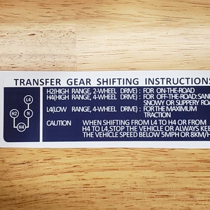 May include: A blue and white transfer gear shifting instructions sticker with a diagram of the gear positions. The text reads: "H2(High Range, 2-Wheel Drive): For On-The-Road, H4(High Range, 4-Wheel Drive): For Off-The-Road: Sandy, Snowy or Slippery Road, L4(Low Range, 4-Wheel Drive): For The Maximum Traction, Caution: When Shifting From L4 To H4 Or From H4 To L4, Stop The Vehicle Or Always Keep The Vehicle Speed Below 5MPH Or 8KM/H."
